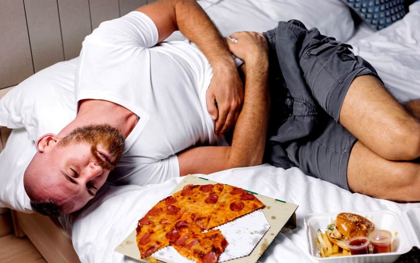 Homem dorme com a mão na barriga cercado por alimentos pesados